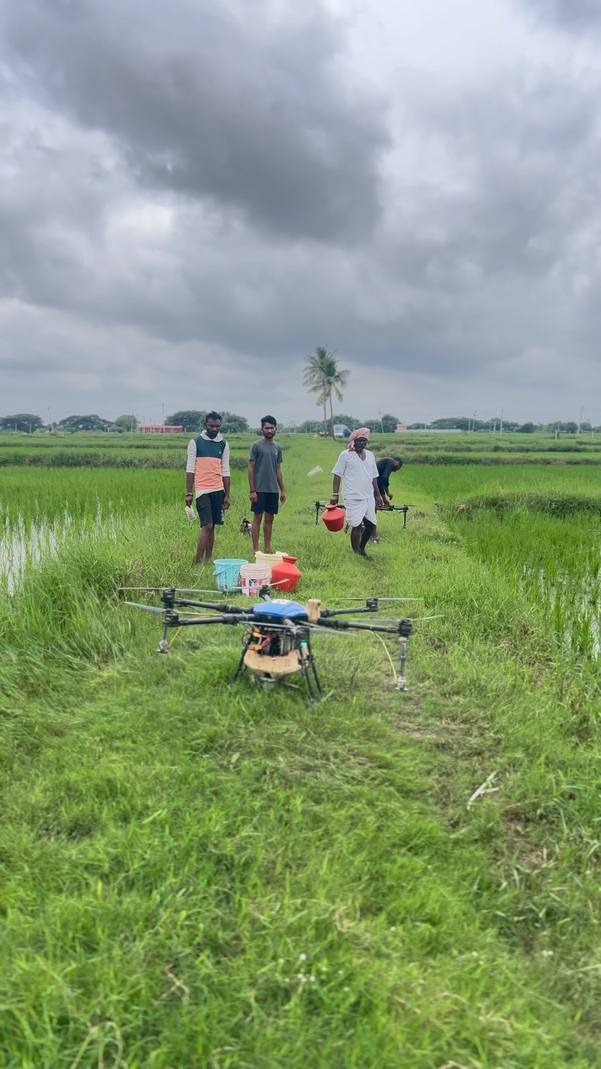 A spraying drone on a bund between paddy fields, with farmers and mixing containers behind it