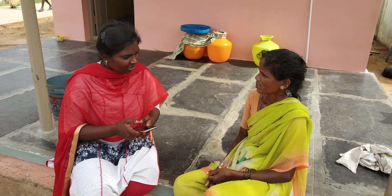 A field surveyor recording a farmer’s responses on a mobile phone while seated with her outside a home in Ananthapur